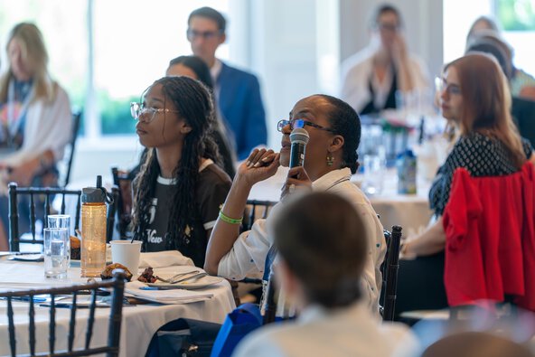 A woman seated at a table holds a microphone waiting to ask a question at an event.