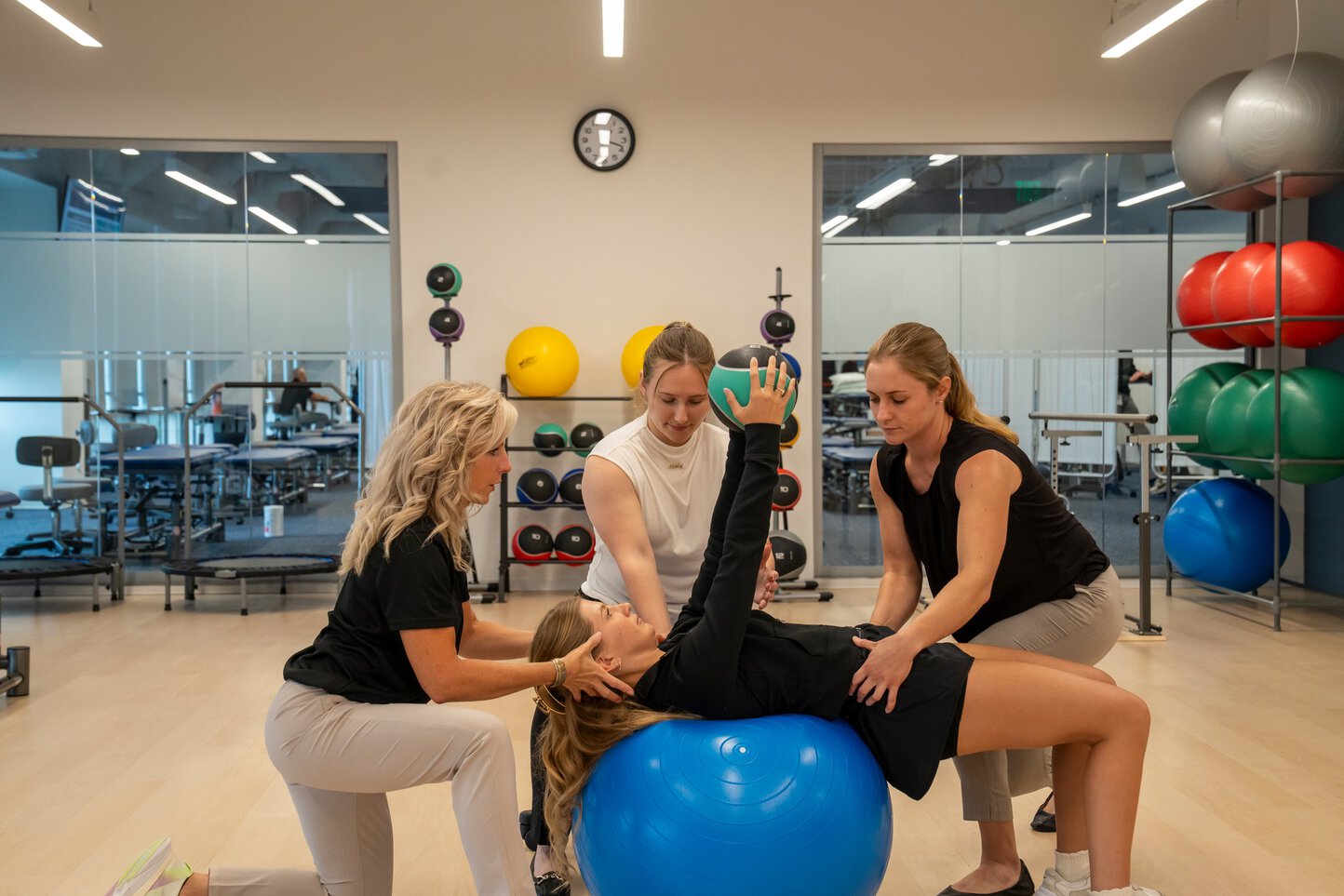 A woman guides three female students through a physical therapy exercise using fitness balls.