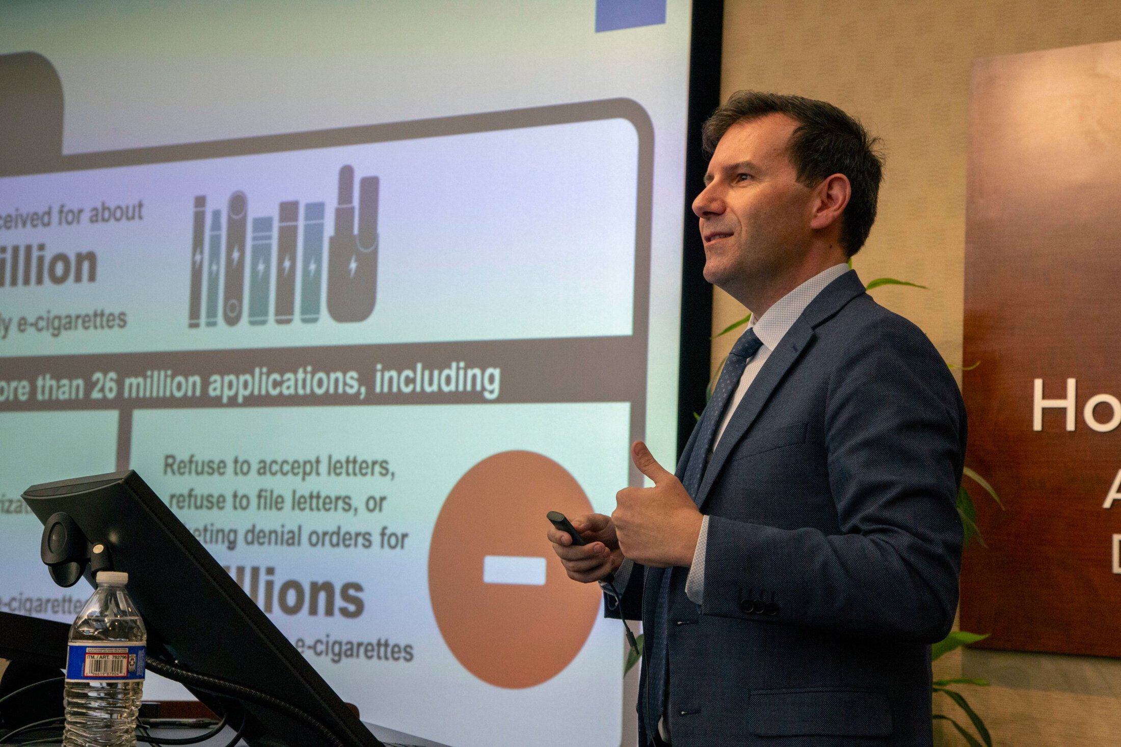 A man speaks at a podium with a large screen behind him showing slides.