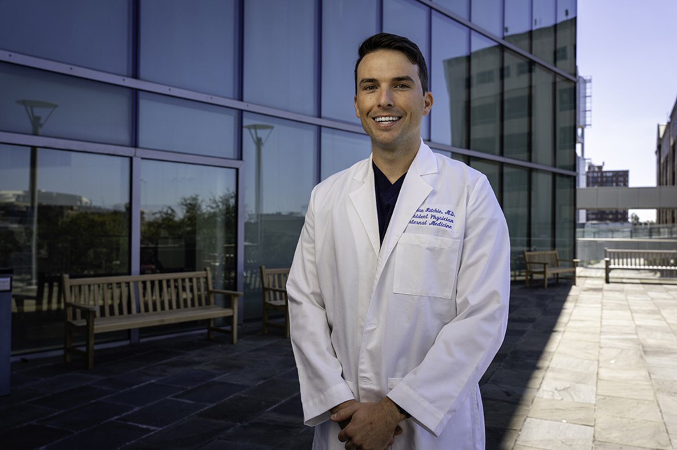 A doctor in a white coat poses in front of a glass-walled building.