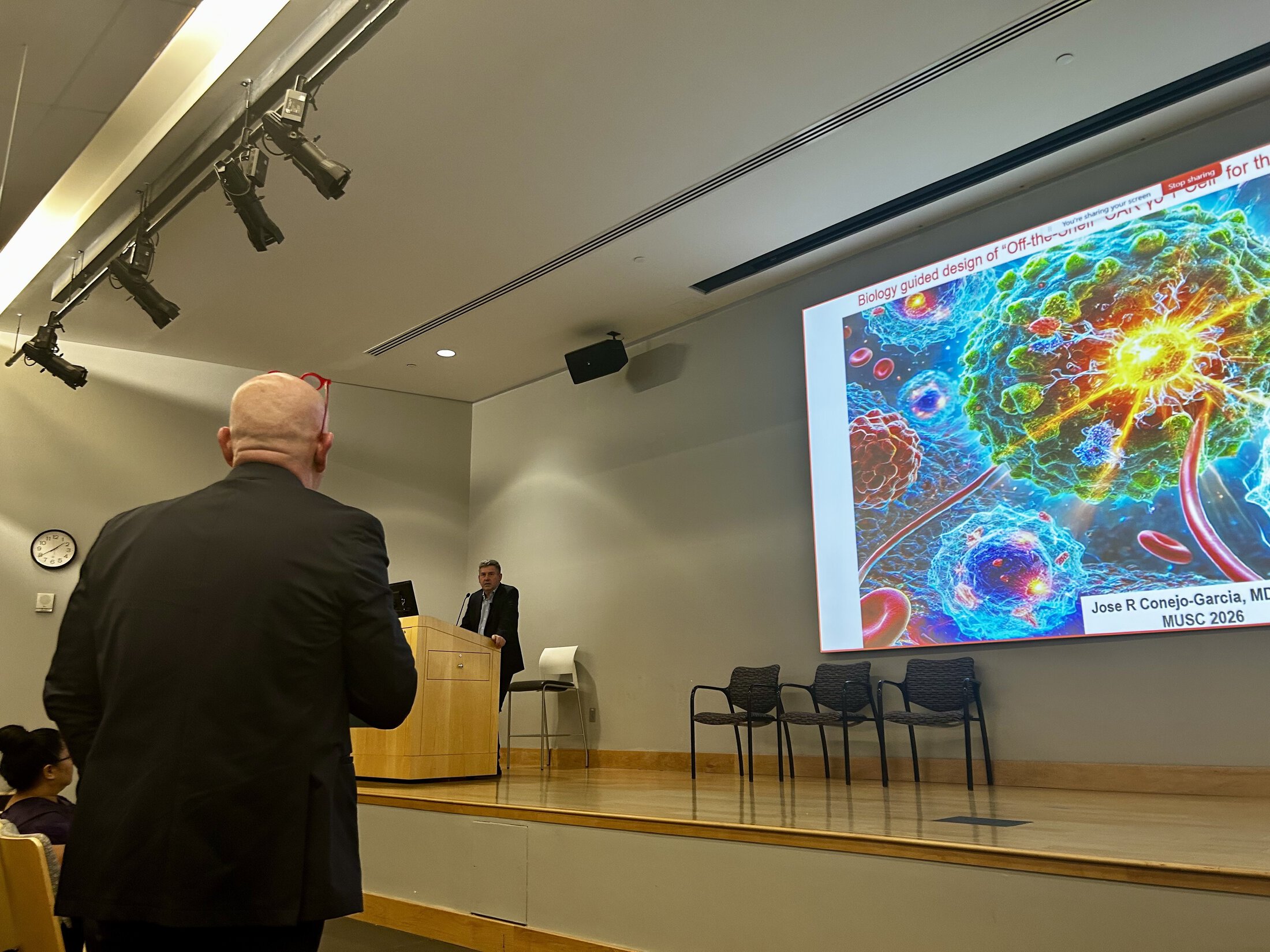 An audience member stands to ask a question during a presentation in a lecture hall, while a speaker stands at a podium on stage. A large screen displays a colorful scientific slide about cellular therapy with detailed imagery of cells.