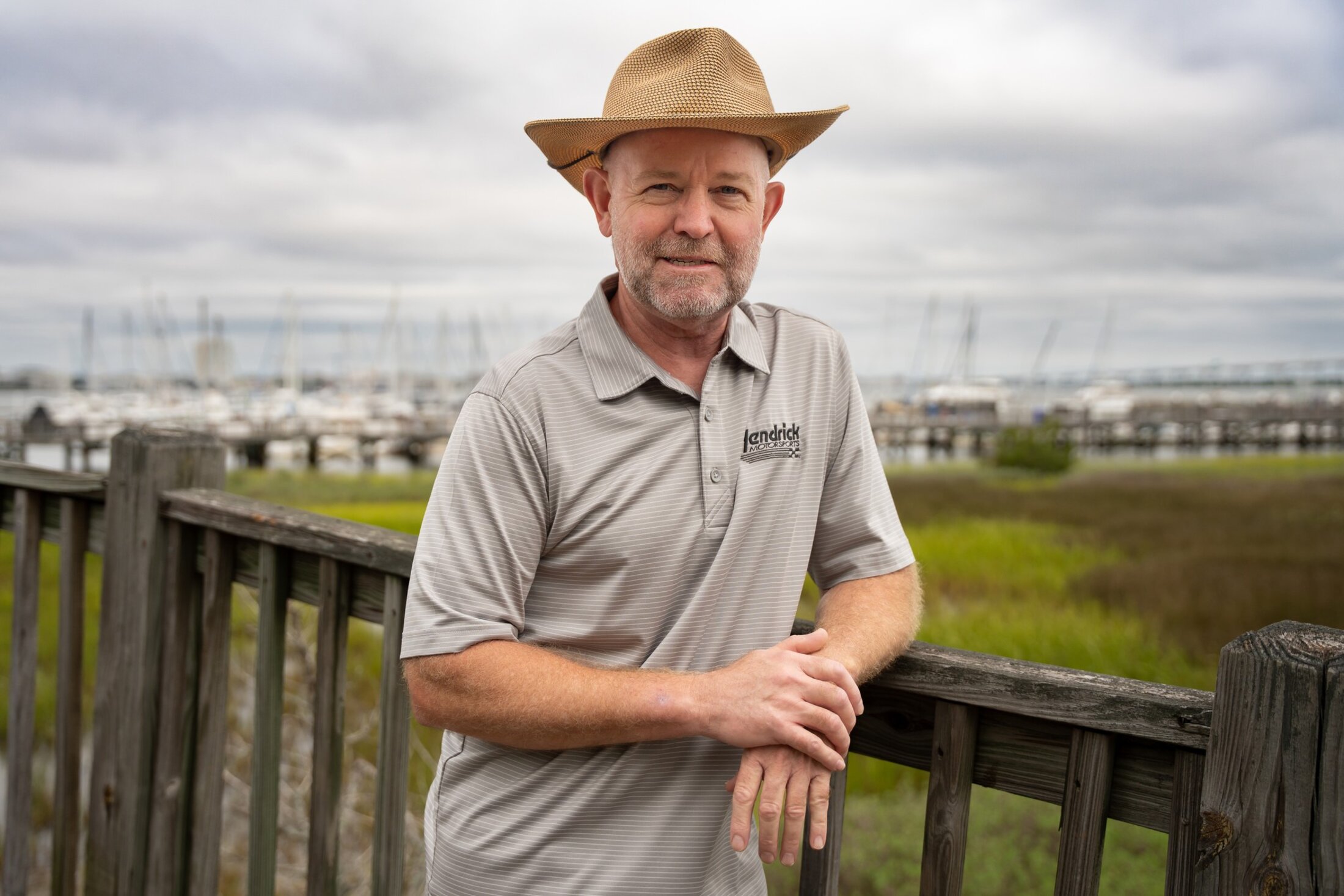 A man in a straw hat poses casually against the fence with the marsh and marina in the background.