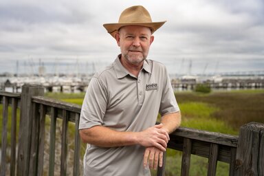 A man in a straw hat poses casually against the fence with the marsh and marina in the background.
