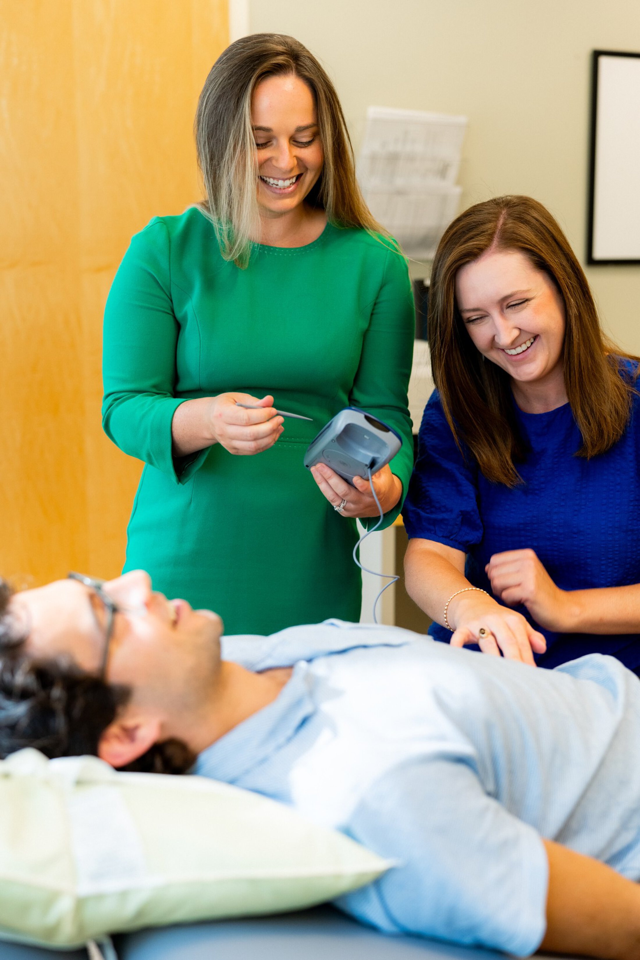 Two women are leaning over a bed where a man with dark hair, glasses and a light blue shirt is lying. They are all smiling. The woman on the left has light brown hair, is wearing a green dress and is holding a device about the size of a large cell phone in one and and a matching stick in the other. The other woman has brown hair, is wearing a blue shirt and may be touching the man's side.