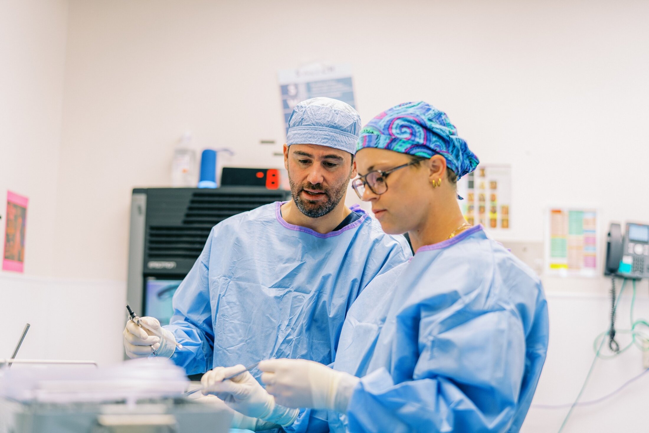 Two urologists in surgeon gowns and caps discuss the surgical tools they are holding.