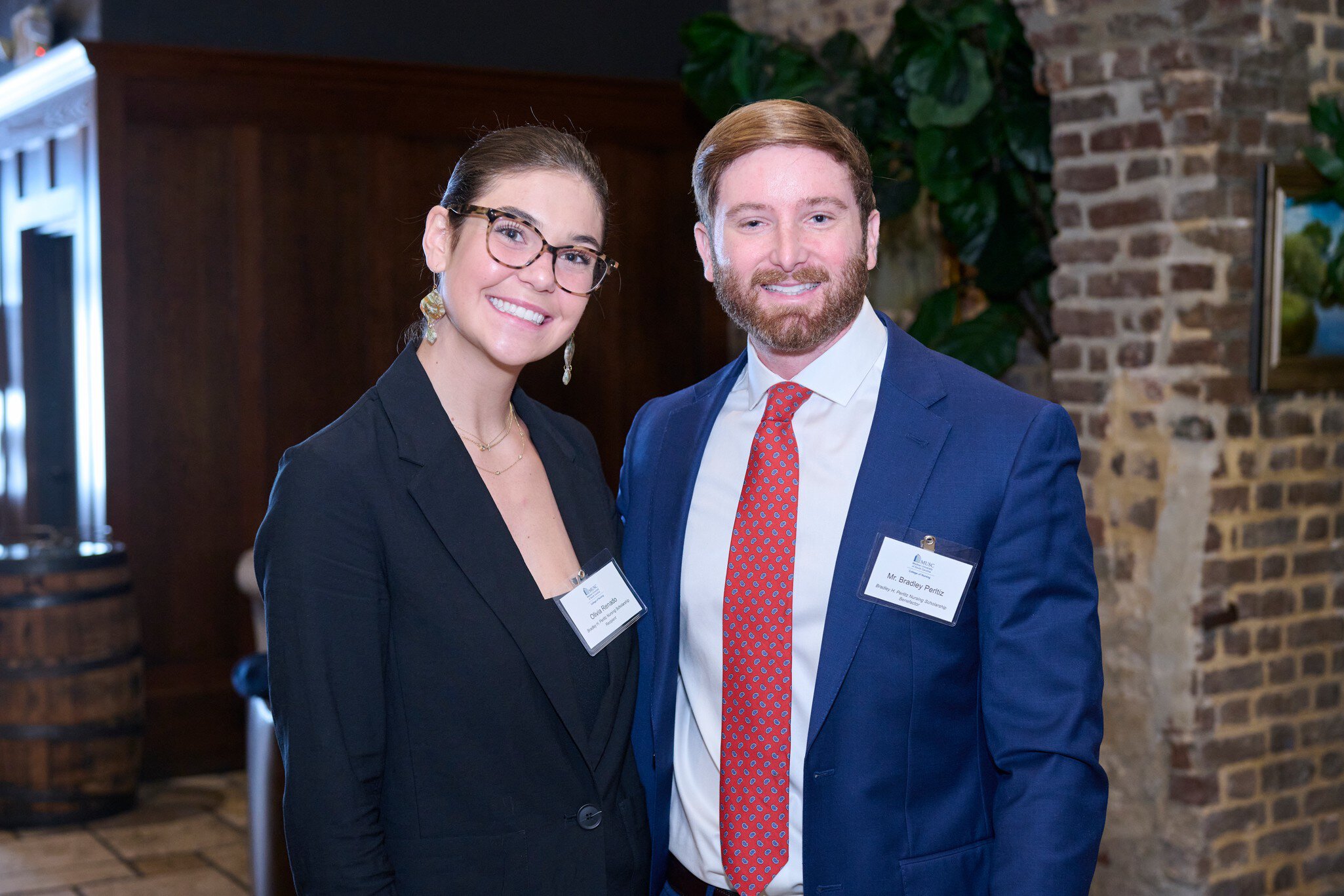 A woman and a man in business attire smile at the camera. She wears glasses and a black blazer, he wears a blue suit and red tie. They stand by a brick wall, conveying a professional and friendly atmosphere.