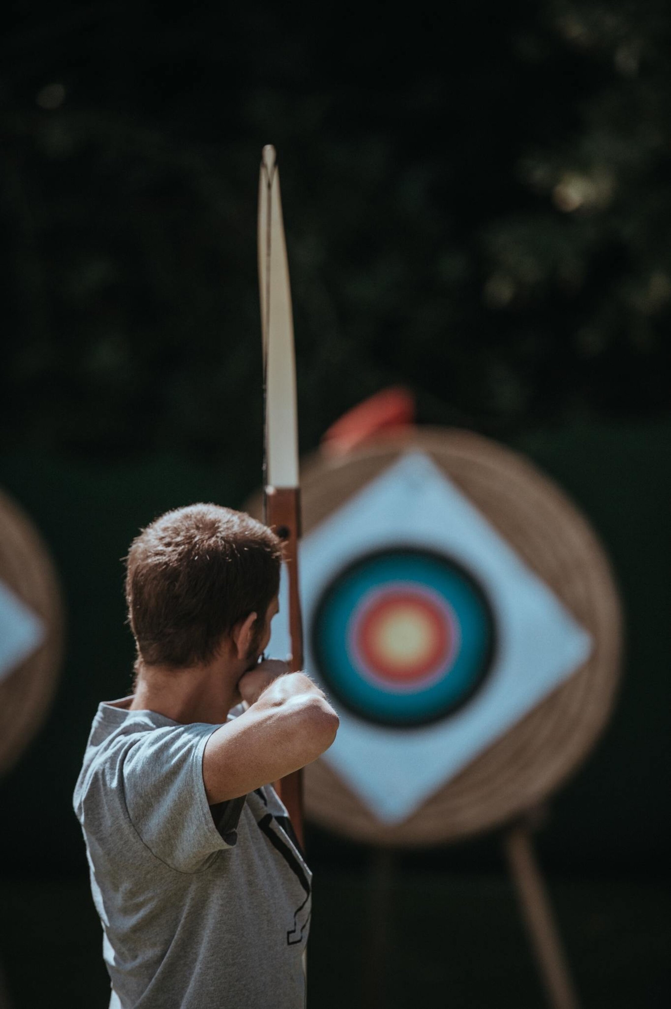 Person drawing a bow and aiming at an archery target outdoors, with the target blurred in the background and the archer in focus.