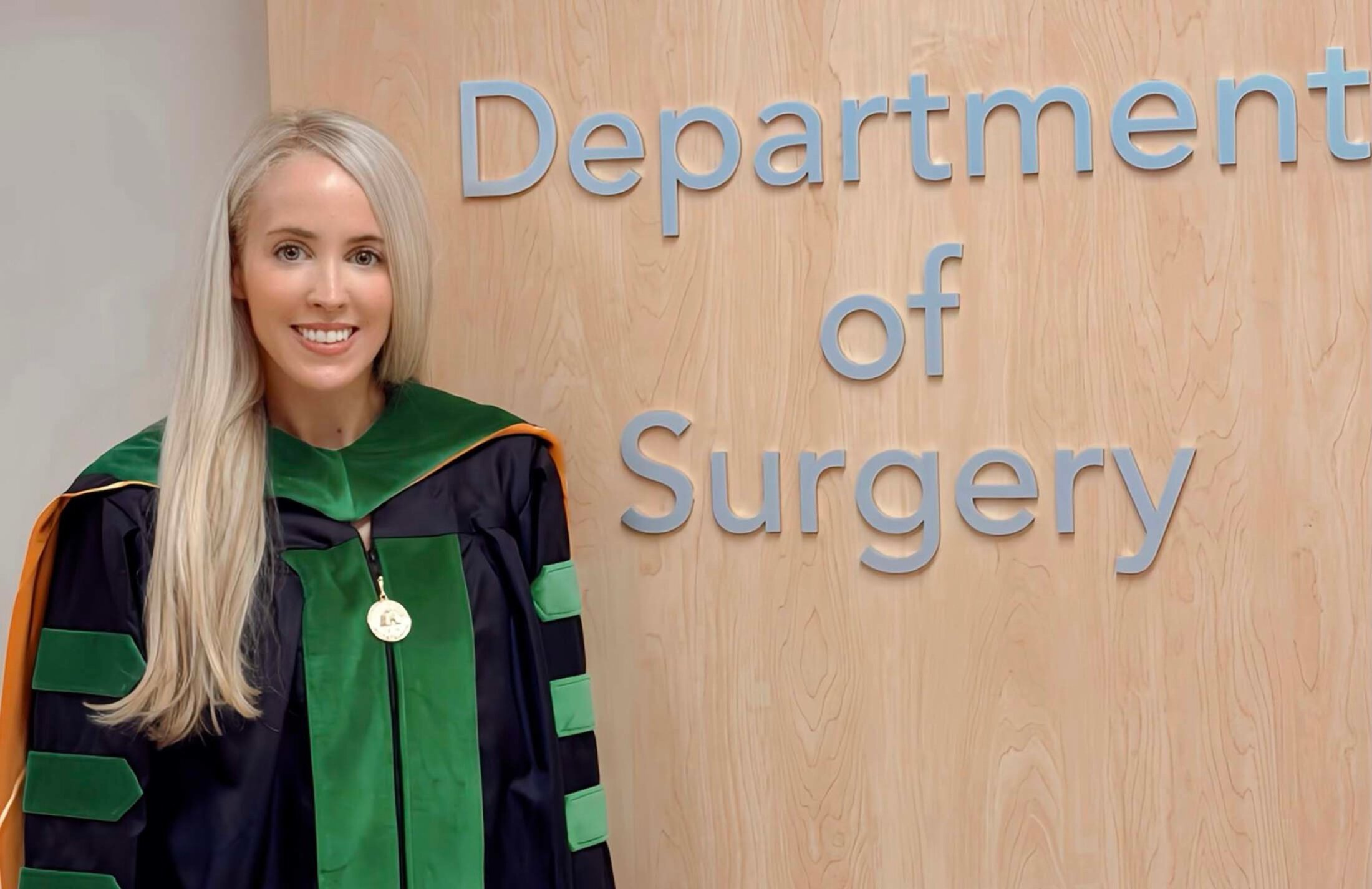 A woman with long blonde hair stands beside a sign that says Department of Surgery. She is wearing a black graduation robe with green accents.
