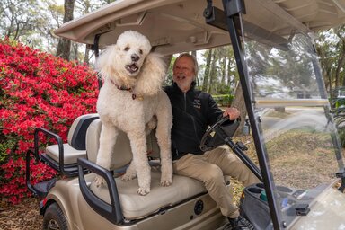 A large white poodle stands eagerly on the seat of a golf cart with her owner in the driver's seat, beaming at her.
