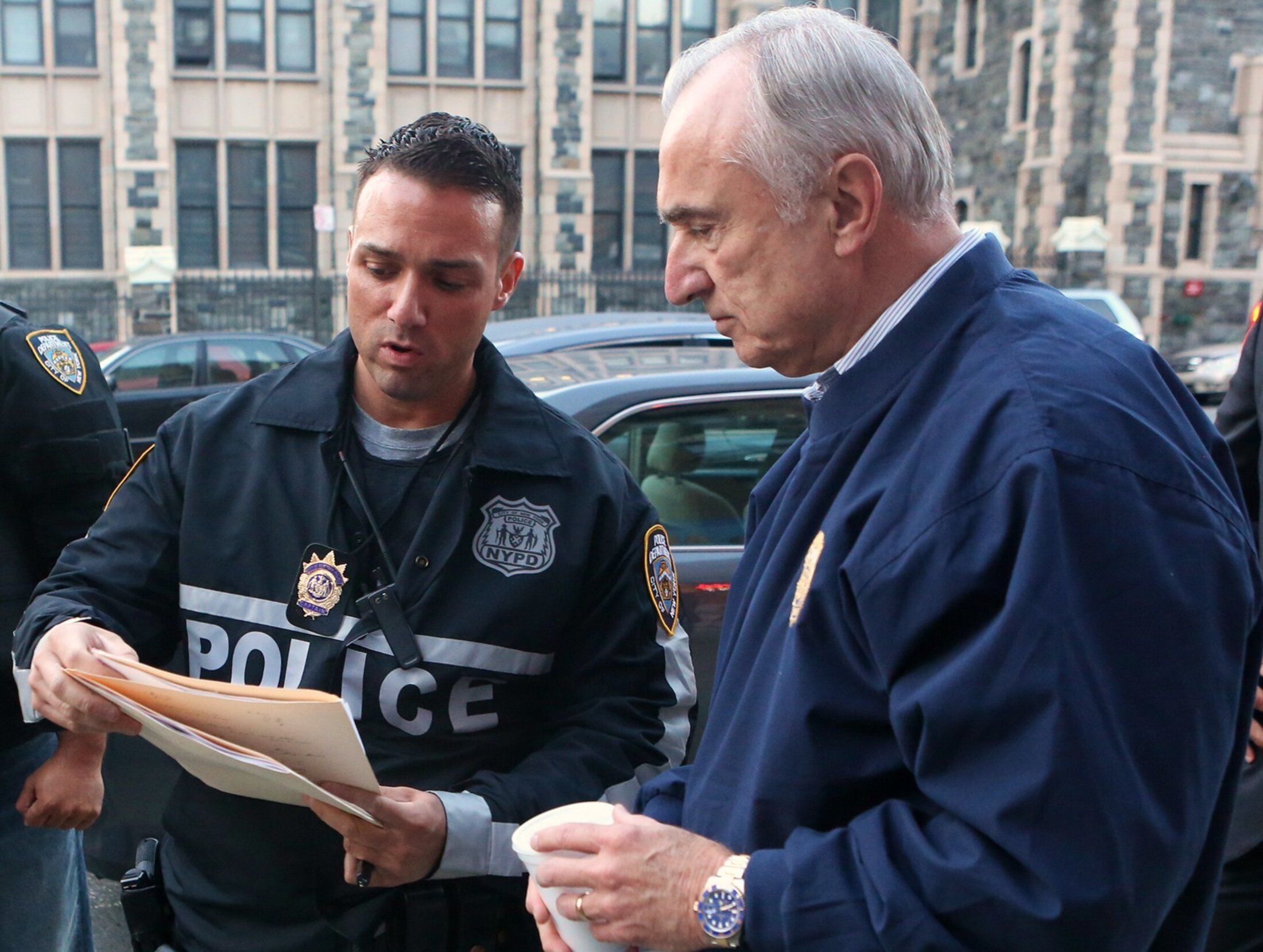 A man wearing an NYPD jacket that says POLICE on it holds a folder as he shows it to a second man who is wearing a navy top with a badge on it.