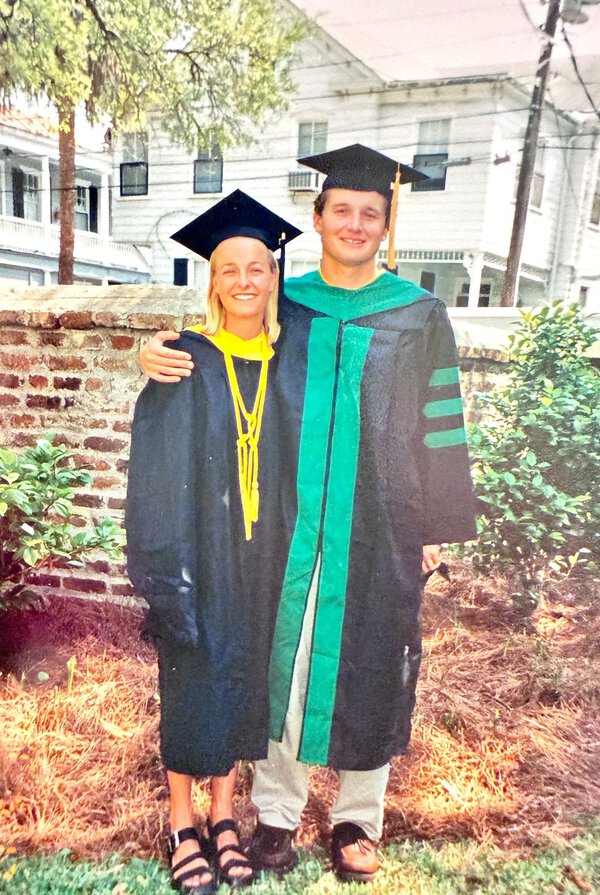 Two people stand in cap and gown together in front of a brick wall.