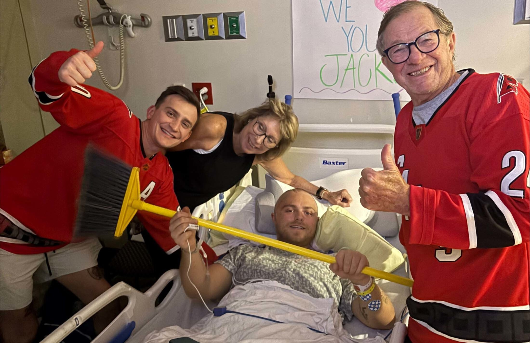 Four people in a hospital room. A young man wit a shaved head is lying the bed holding a broom. A young man in a red jersey is leaning in next to him with his thumb raised. A woman with light hair and eye glasses is leaning in with her arm on the bed. A man wearing a red jersey stands on the right giving a thumbs up.