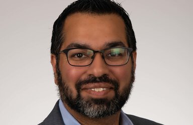 Headshot of a man with dark hair, a beard and black-framed eyeglasses. He is wearing a blue shirt under a dark gray jacket and smiling.