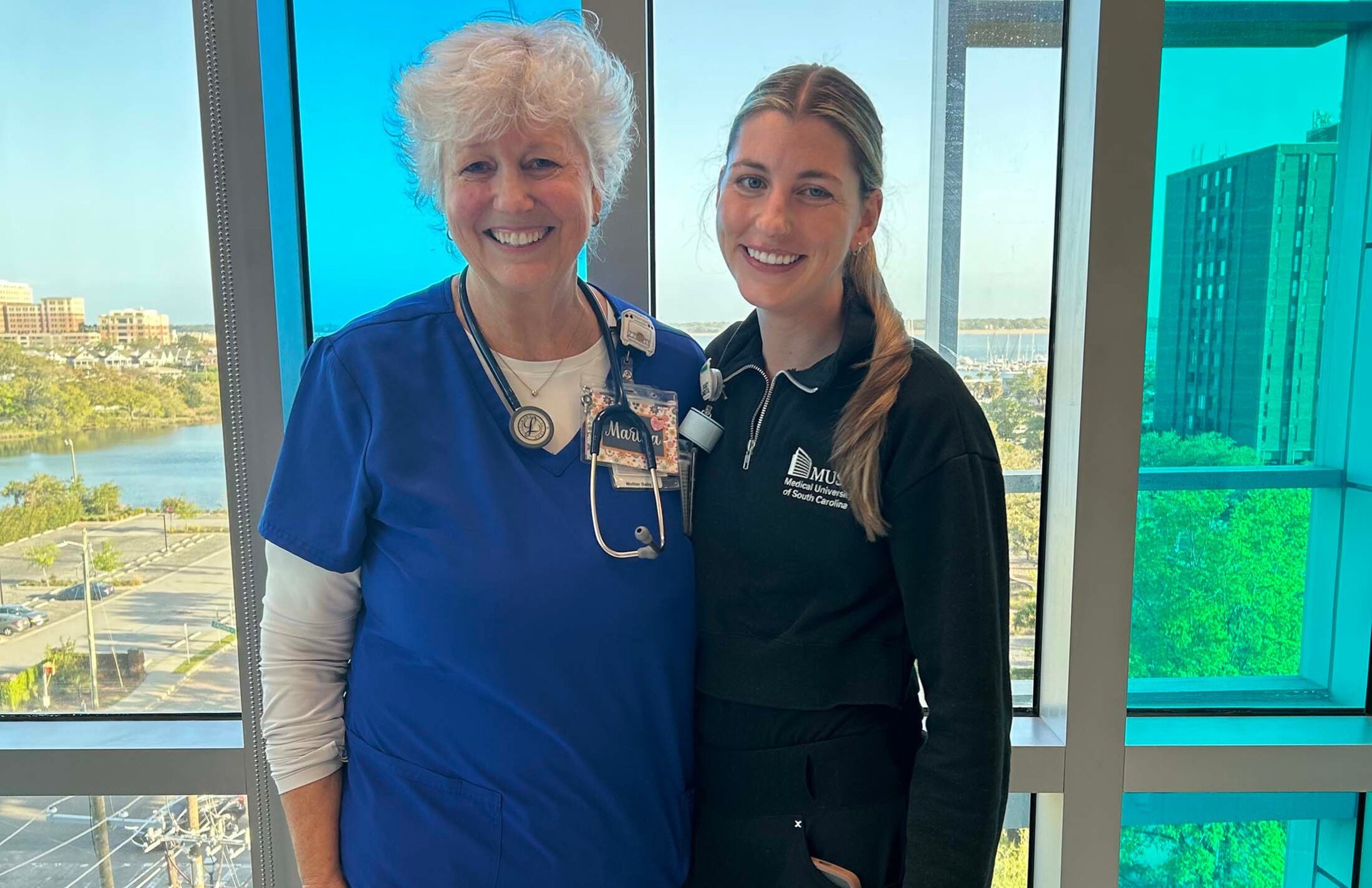 Two women stand in front of a large window. The woman on the left is wearing royal blue scrubs. The woman on teh right is wearing a dark jacket and pants. She has a ponytail.