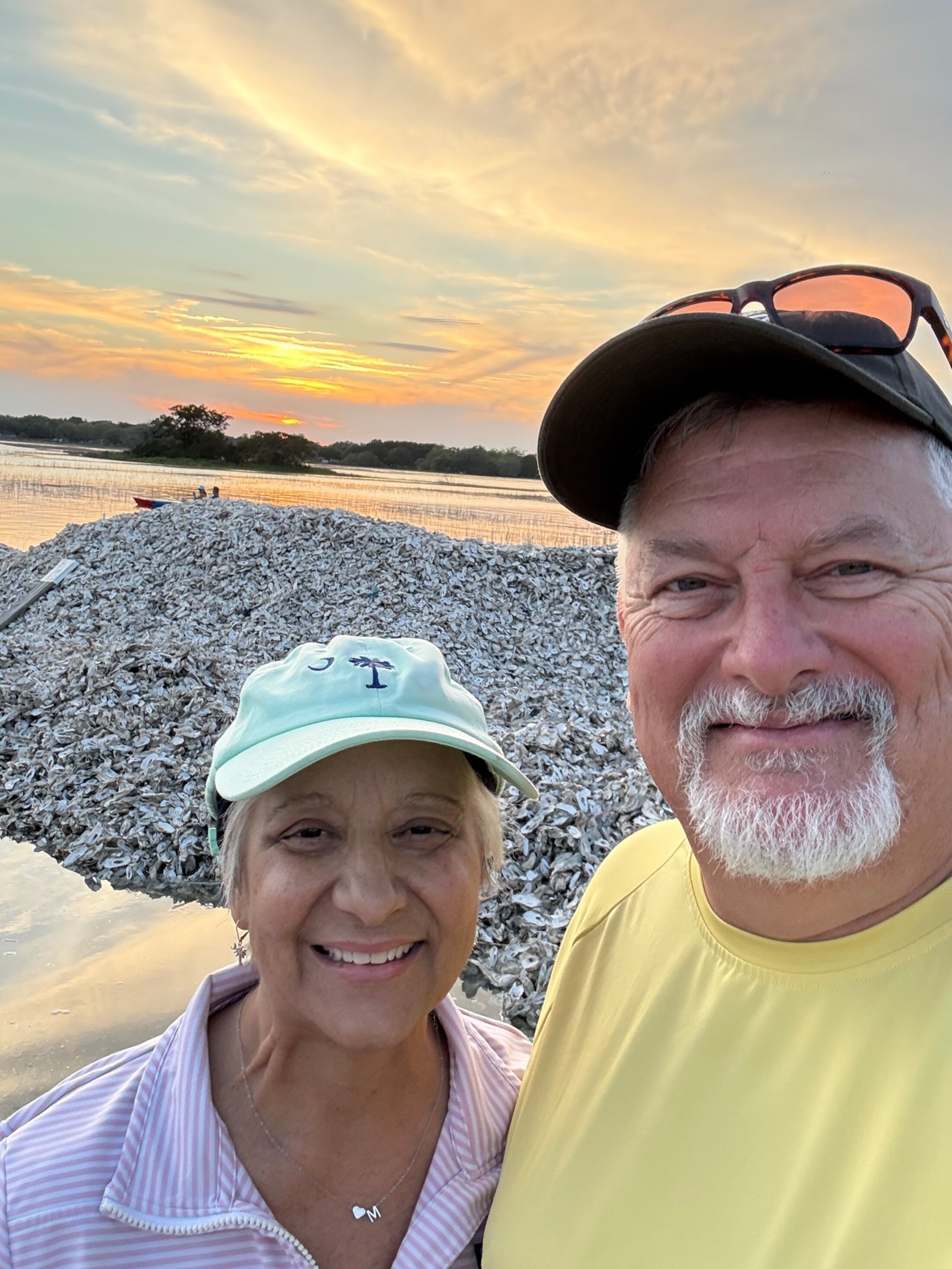 An older couple pose for a selfie at at the beach with oyster shells and kayakers in the background.