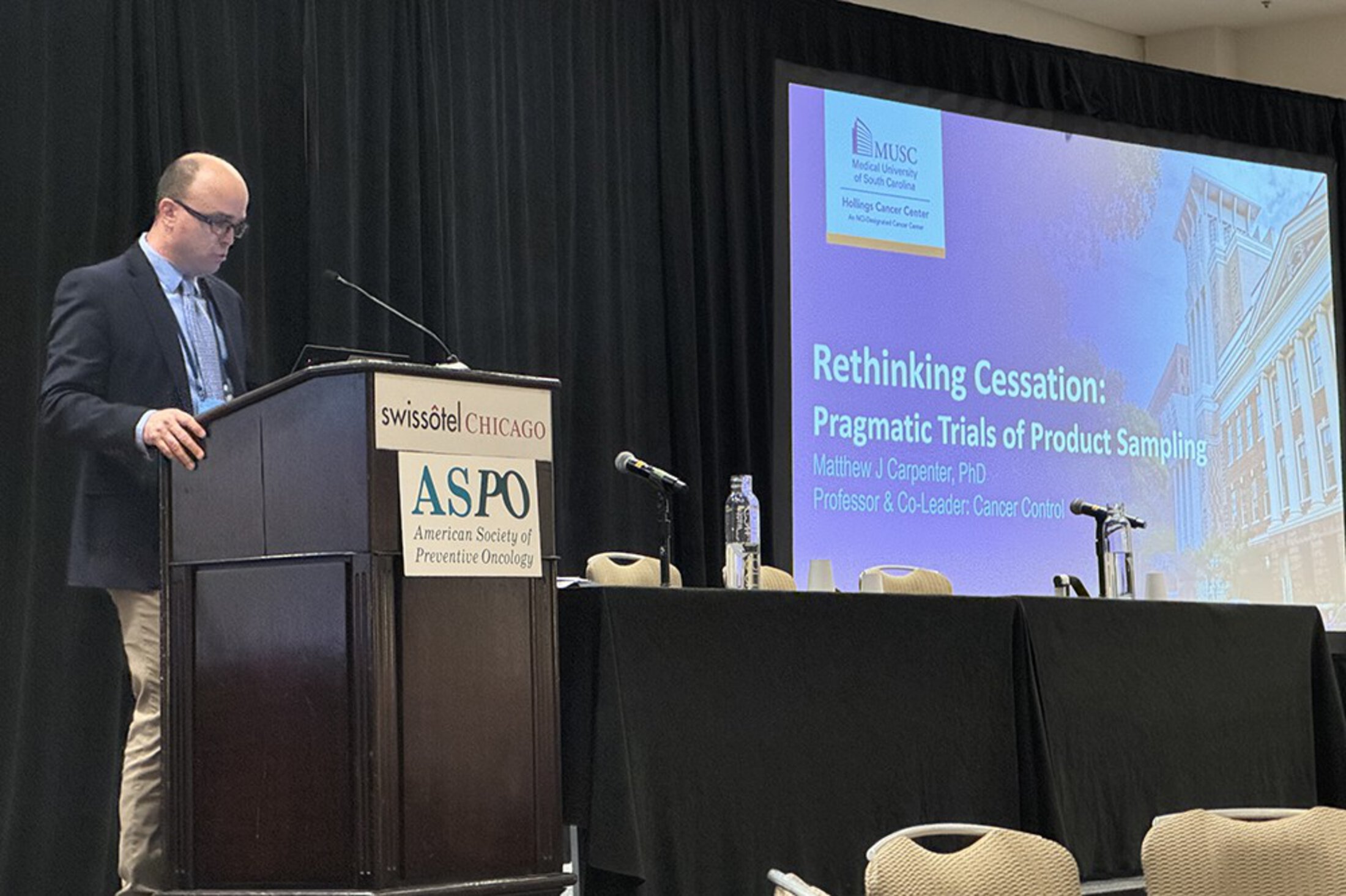 A man stands at a podium. A large screen behind him displays an introductory slide that says Rethinking Cessation: Pragmatic Trials of Product Sampling. Matthew Carpenter, Ph.D.