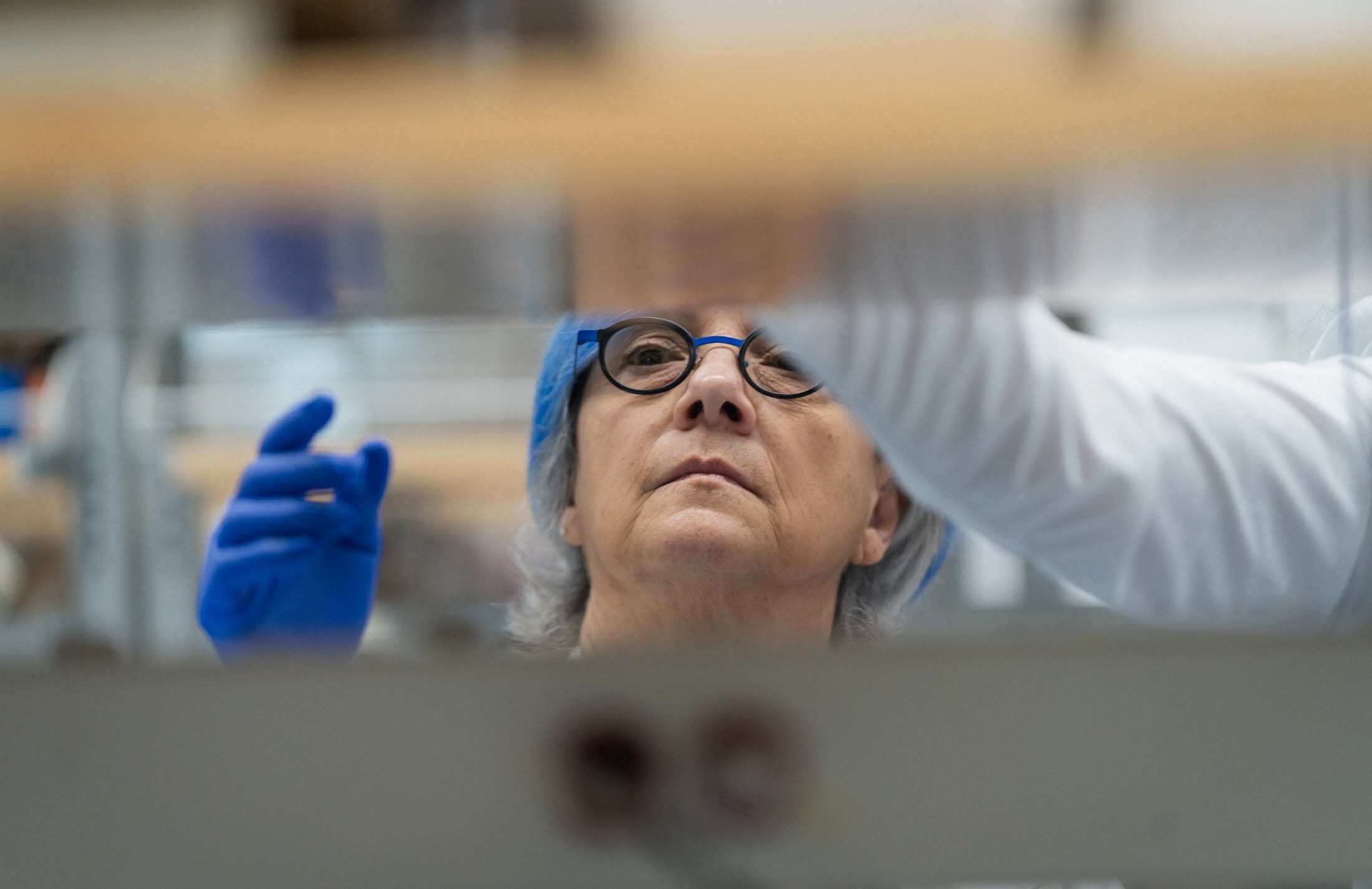 Woman's face through what look like white slates. She has blue and white hair and is wearing glasses. She is also wearing a blue glove.