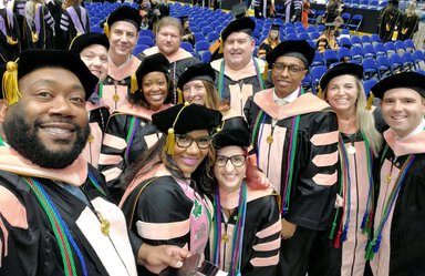 Twelve people in black graduation robes with salmon colored accents smile in a group selfie. The camera appears to be held by the man on the far left. They are smiling, standing in front of a rield of royal blue chairs.