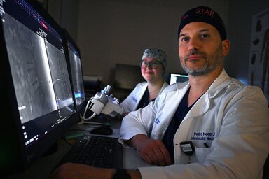A man wearing a head covering looks at the camera. He is wearing a white coat with the name Pedro Norat embroidered on the left. A woman is seated beside him, also looking at the camera. They are sitting in front of computer monitors.