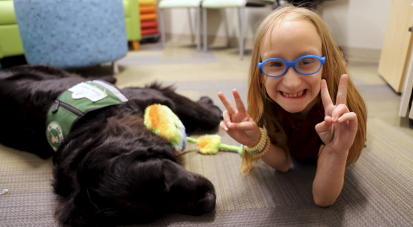 A child patient holding up a peace sign while having a therapy session with a black lab dog