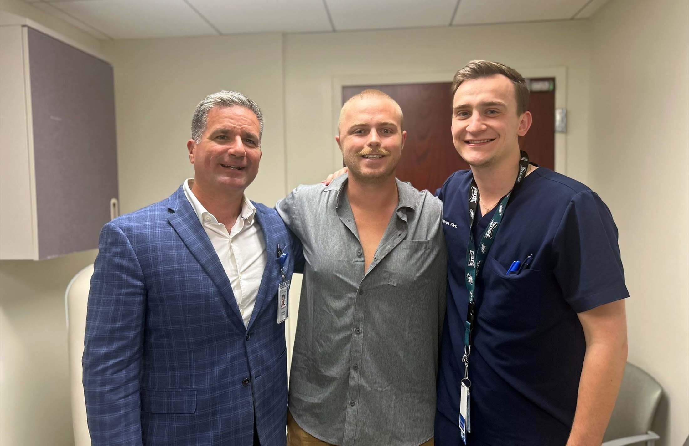 Three men stand together. The one on the left has gray hair and is wearing a white shirt and blue checked coat. The one in the middle has short blond hair and is wearing a gray shirt. The man on the right is wearing blue scrubs and a lanyard with a tag o nit.