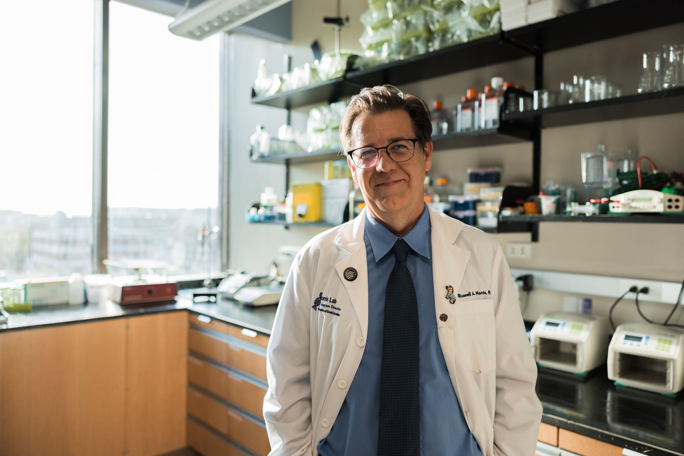 A man wearing a white lab coat, blue shirt and necktie smiles as he stands in front of shelves filled with glass containers. There is a large window on the left side of the photo.
