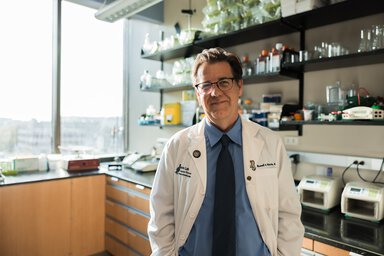 A man wearing a white lab coat, blue shirt and necktie smiles as he stands in front of shelves filled with glass containers. There is a large window on the left side of the photo.