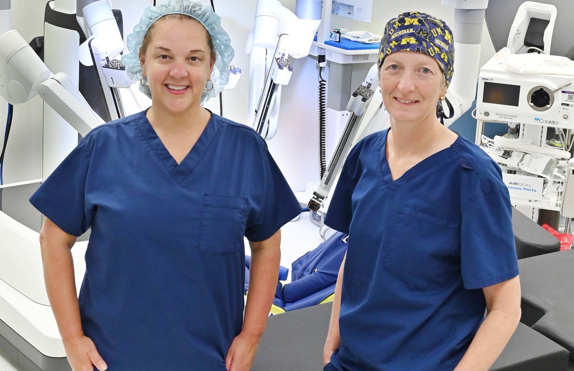 Two women wearing headcovers and blue scrubs smile while standing in front of a white machine with multiple arms.