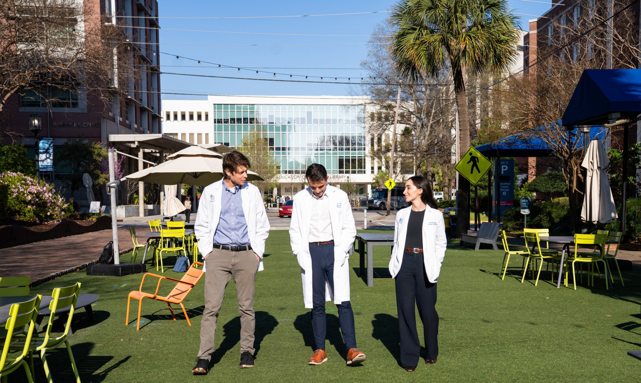 Three young people in medical white coats walk along a pedestrian gathering space on an urban medical university campus.