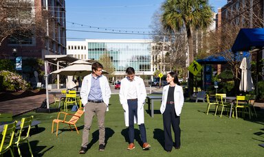 Three young people in medical white coats walk along a pedestrian gathering space on an urban medical university campus.