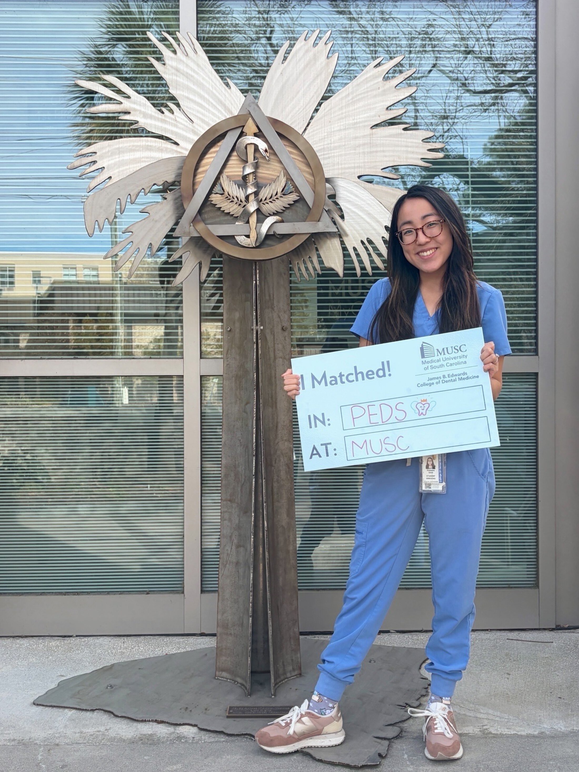 Woman in blue scrubs smiles while holding a “I Matched! In: PEDS, At: MUSC” sign beside a healthcare-themed sculpture outside a university building.