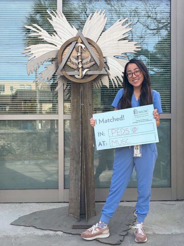 Woman in blue scrubs smiles while holding a “I Matched! In: PEDS, At: MUSC” sign beside a healthcare-themed sculpture outside a university building.