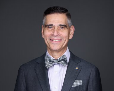 Professional headshot of a smiling man wearing a dark suit jacket and patterned bow tie against a dark background.