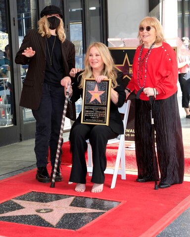 Three women on a red carpet that surrounds a pink star on what looks like black marble. The woman on the left is wearing a hat and a surgical mask and dark clothing. The woman in the middle is seated holding a cane and a plaque with a star and the name Christina Applegate on it. Teh woman on the right also has a cane but is standing.