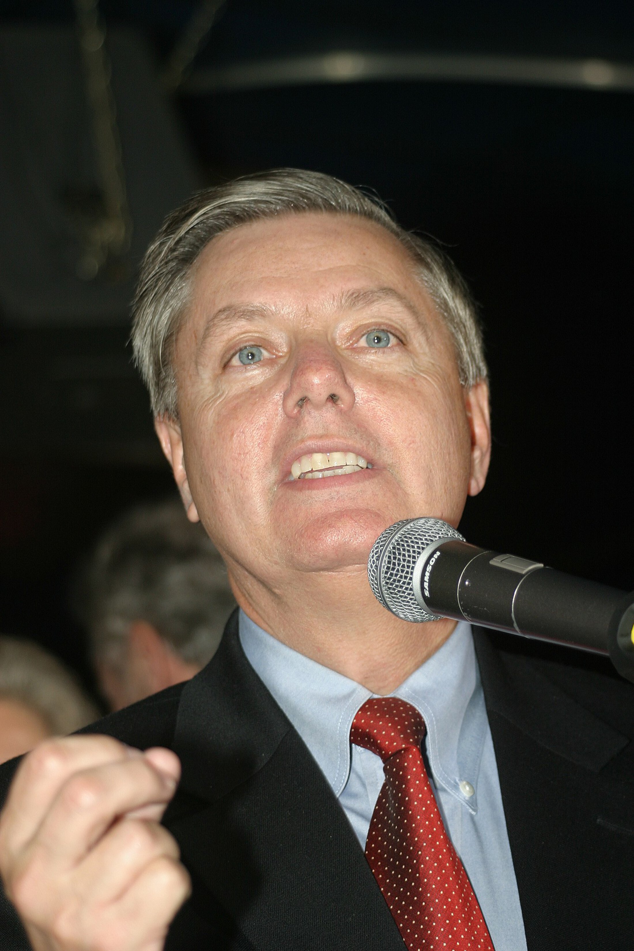 Headshot of a man at a microphone. He has grey/brown hair and is wearing a blue button down shirt, a red tie and a dark suit coat.