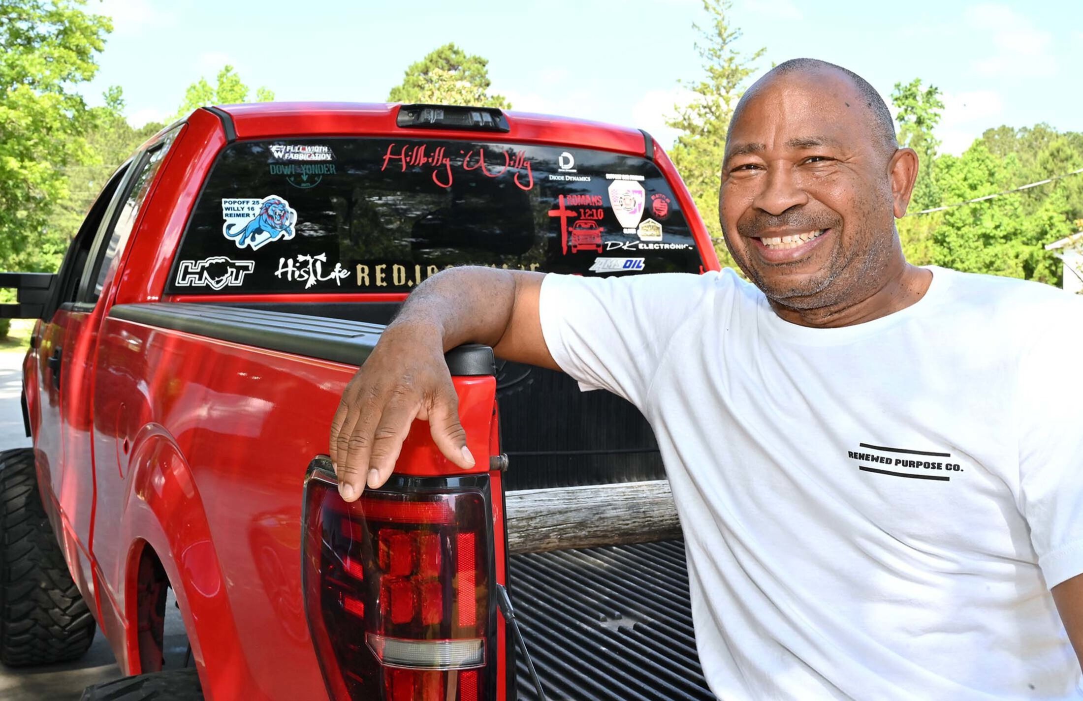 Man wearing t-shirt that says renewed purpose company has his arm up on the bed of a red pickup truck. It has stickers on the window including 