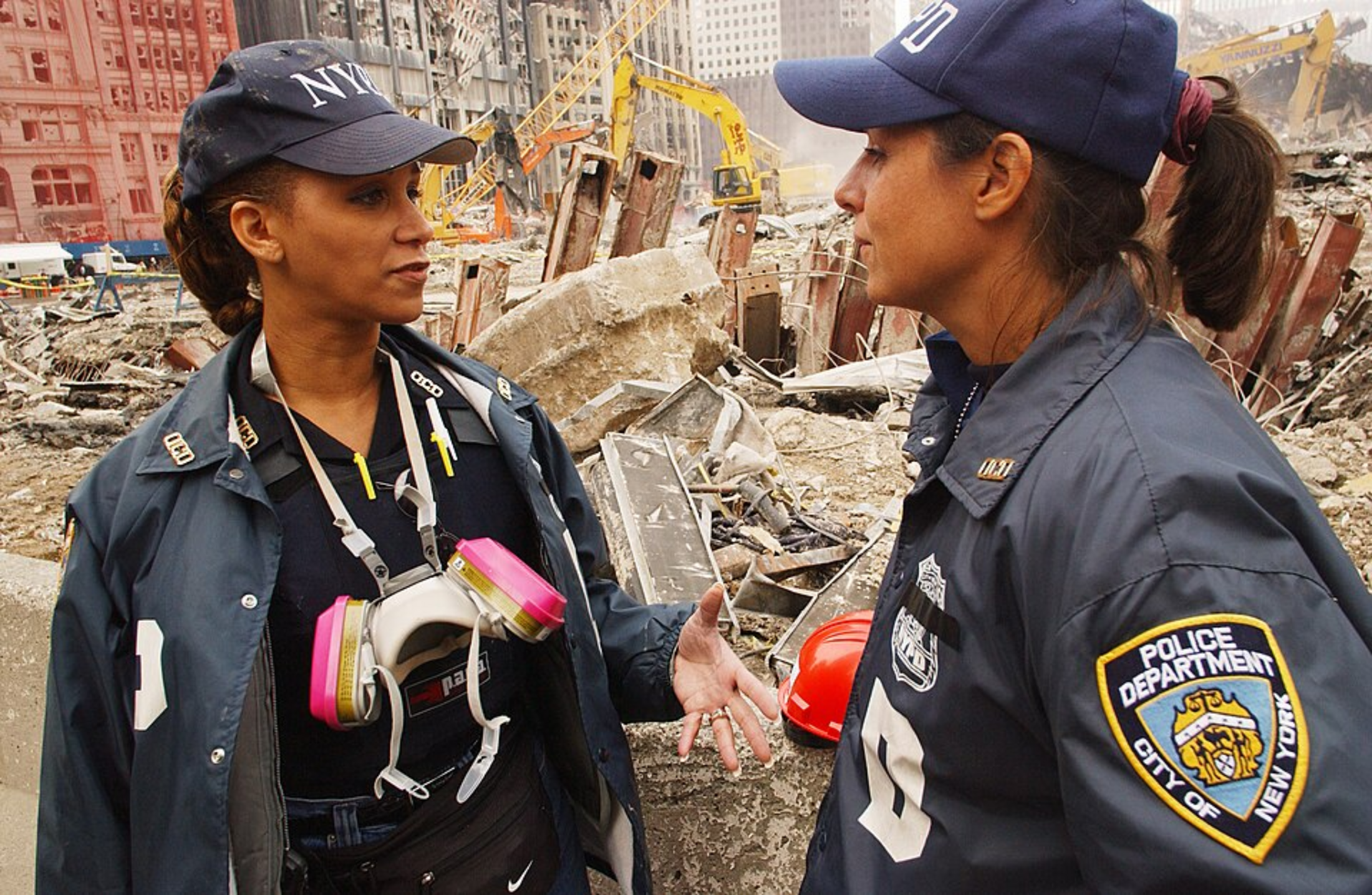 Two women wearing NYPD hats and jackets stand talking in front of a huge pile of debris. One woman has a pink and white mask around her neck.