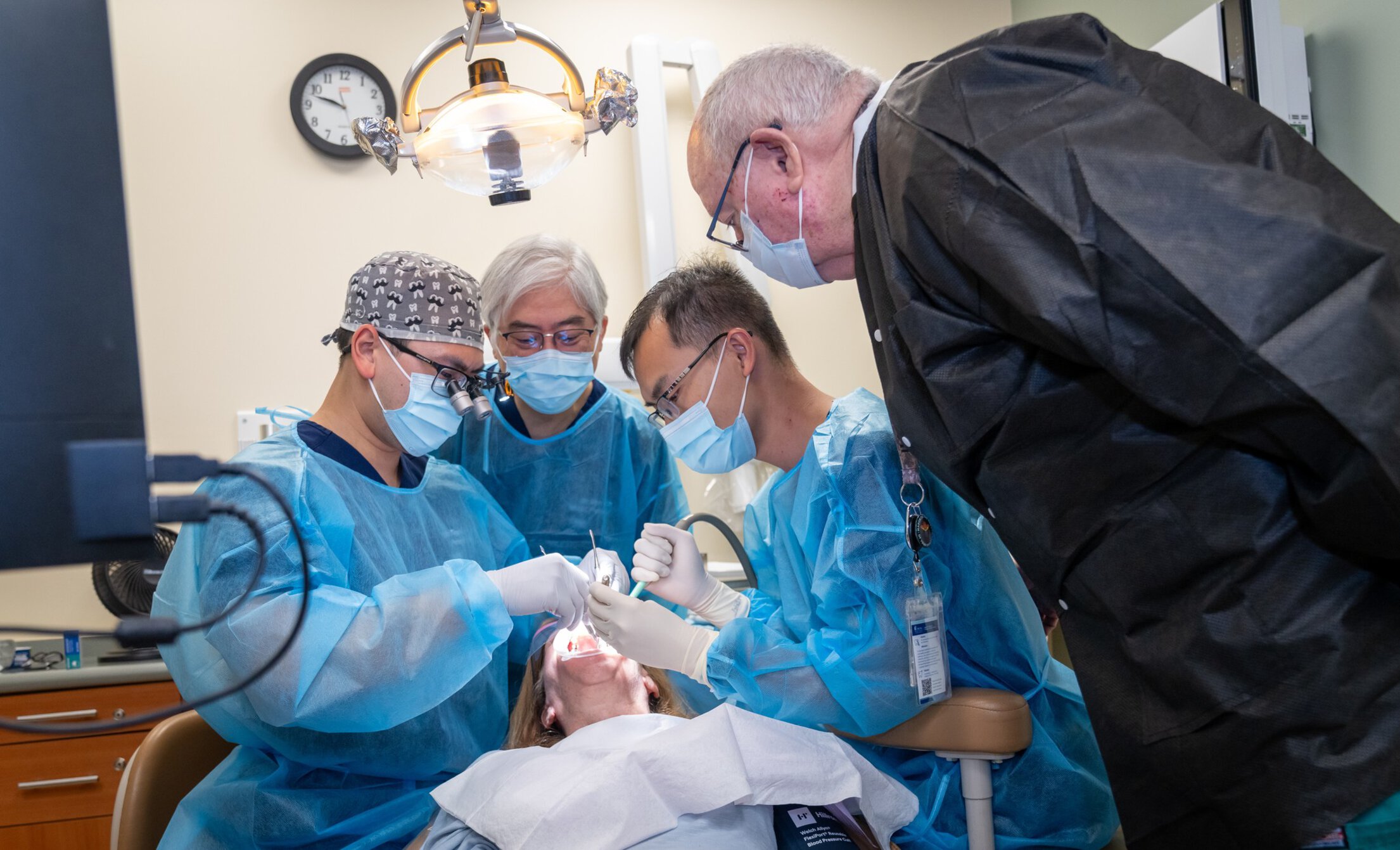 Two periodontics residents examine a patient's mouth. Two faculty periodontists are watching closely.