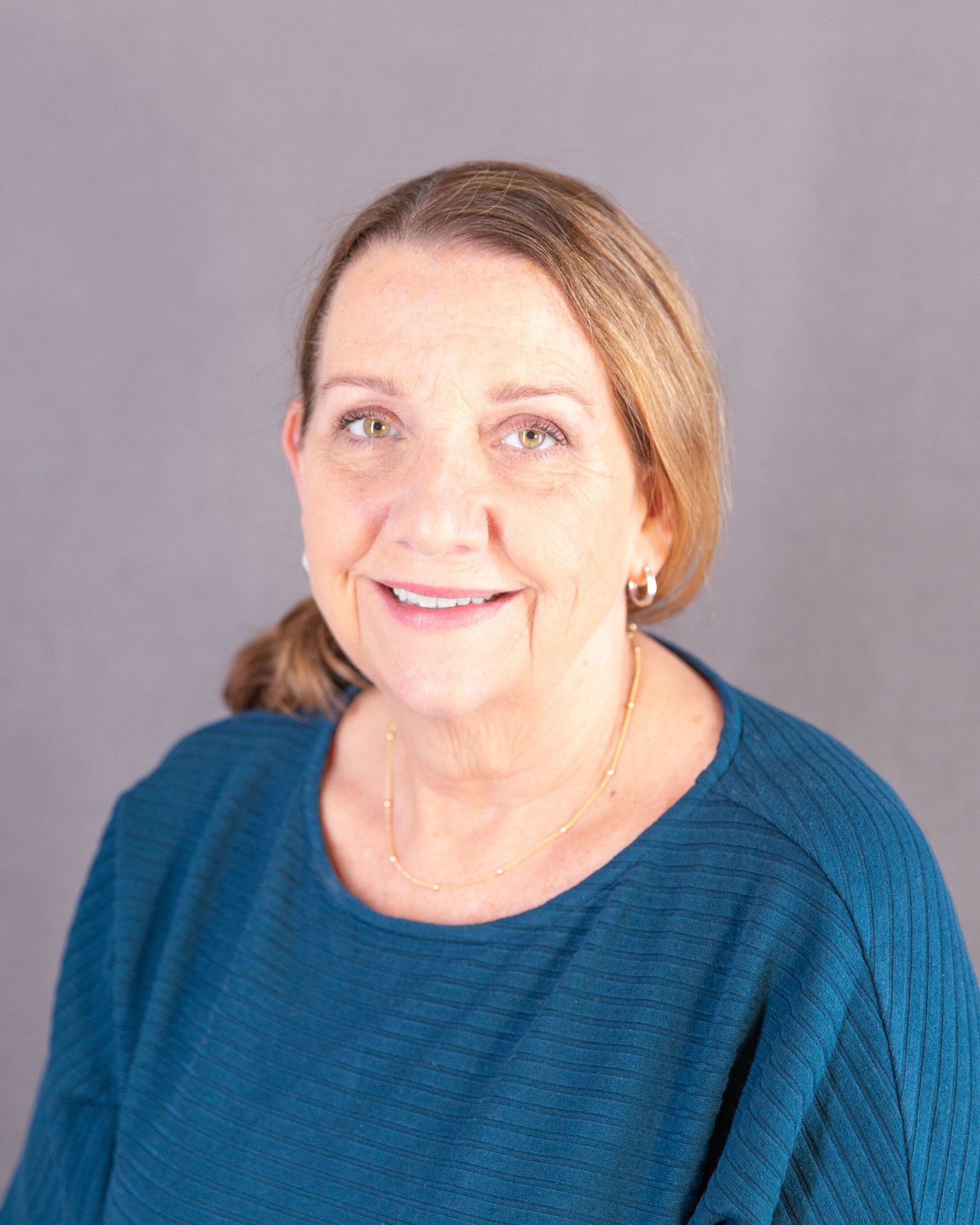 Portrait of a smiling woman with light brown hair pulled back, wearing a teal top and small hoop earrings, against a neutral gray background.