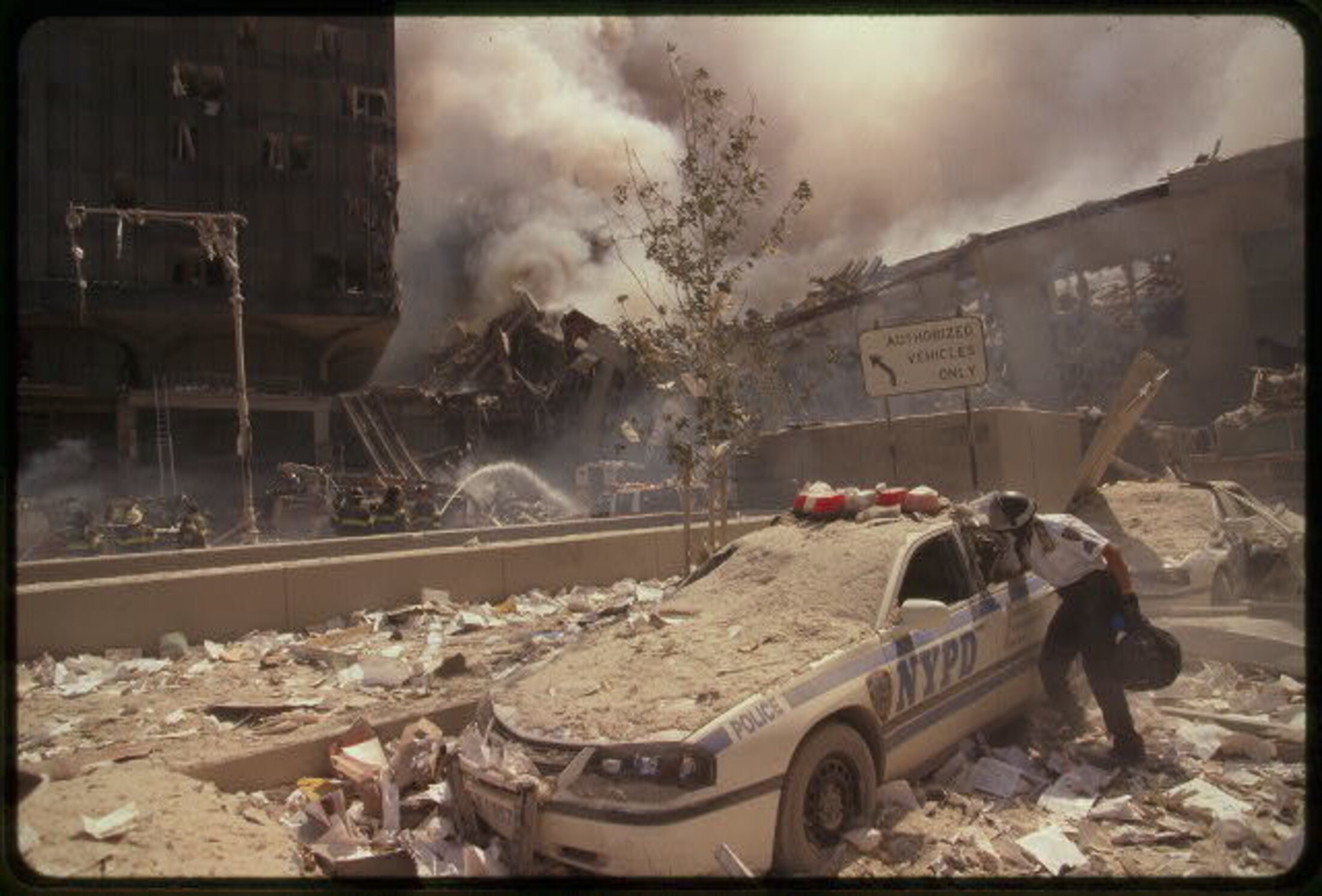 An NYPD police car covered in debris next to a destroyed building. There is smoke rising in the background. A man is reaching into the back seat of the police car.