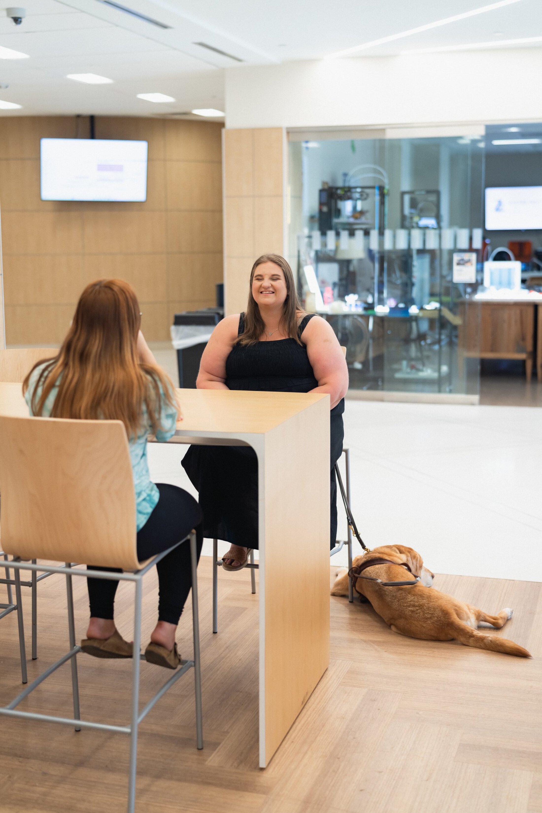 A young woman wearing a black dress is seated at a brown table across from a woman wearing a shirt and pants. The woman in the dress has a reddish colored dog lying on the floor beside her.