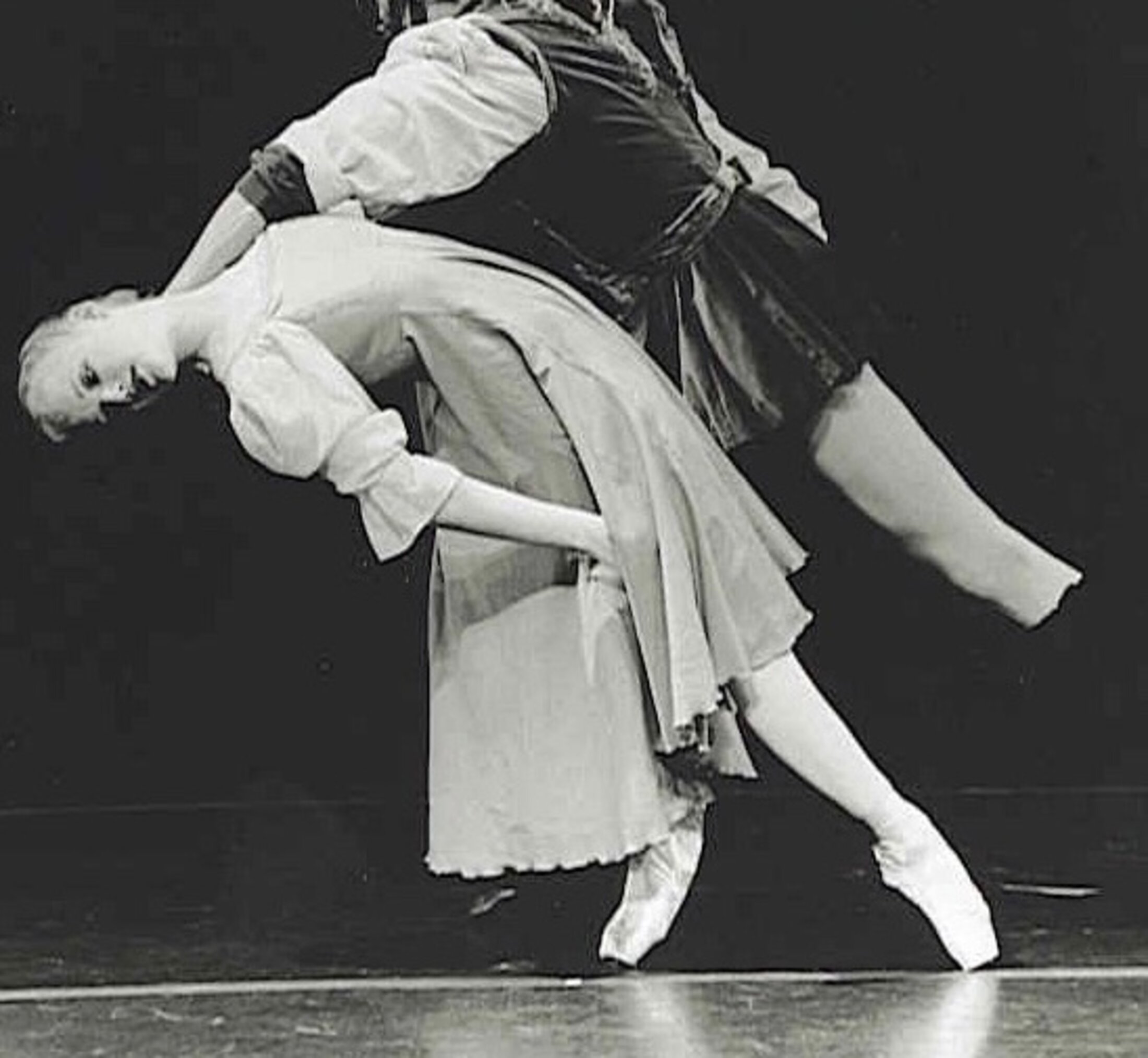 A ballet dancer leans back while a man holds her shoulder. She is wearing a dress and pointe shoes