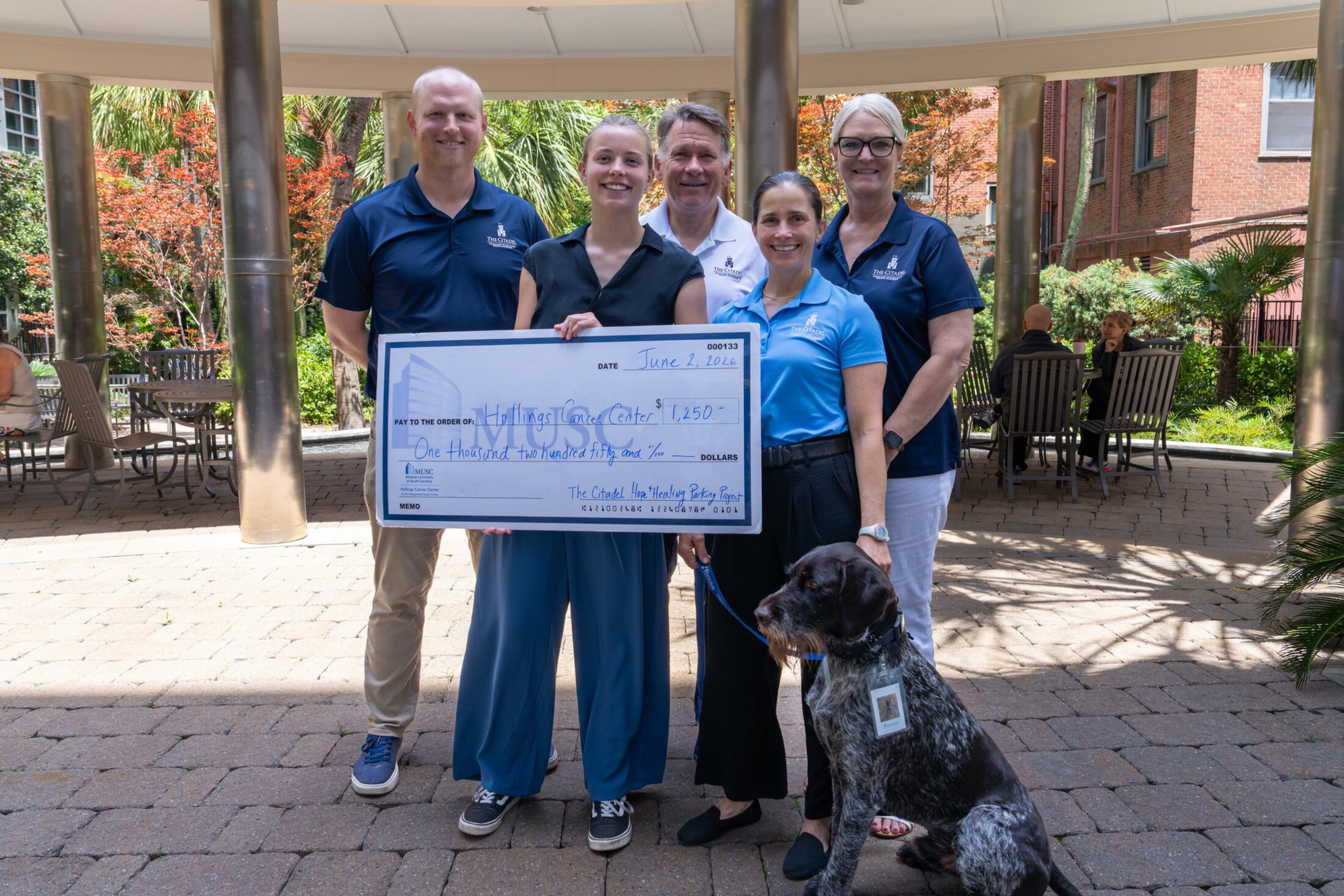 A small group of people and a dog with an oversized check made out to Hollings Cancer Center for $1250.
