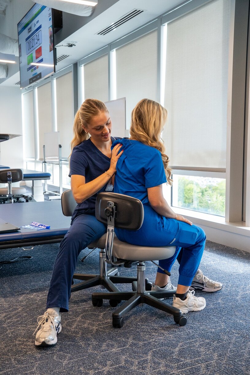 Two students in blue scrubs practice a stand-assist lift in a simulated clinical environment, with one student seated in a chair and the other assisting as part of an OT lab immersion.