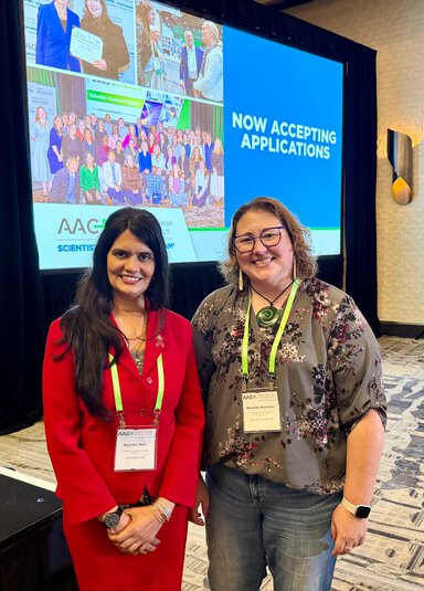 Two women wearing conference lanyards pose in front of a large screen displaying AACR messaging in a hotel conference room.