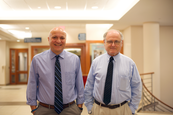 Two men in blue shirts and ties pose in a wide lobby hall.