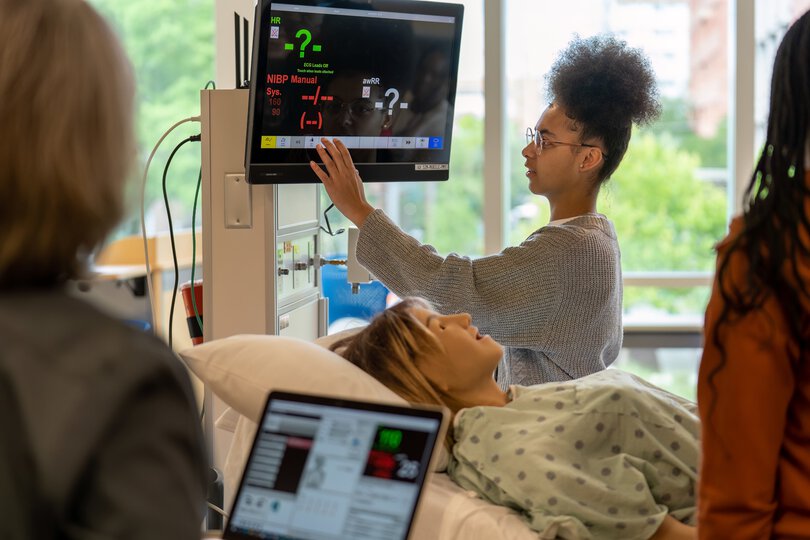 A student interacts with a touchscreen next to a bed with a manikin on it as part of a simulated experience of caring for a patient.