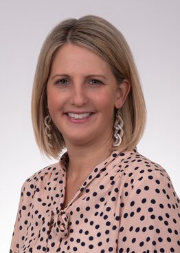 Headshot of woman with shoulder length blonde hair. She has large earrings and a pink and black polka-dot blouse.