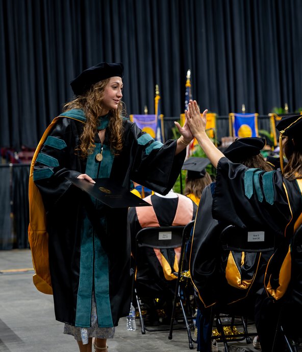A graduate in academic regalia holding a diploma gives a high-five to another graduate during a commencement ceremony on a stage with seated attendees and banners in the background.