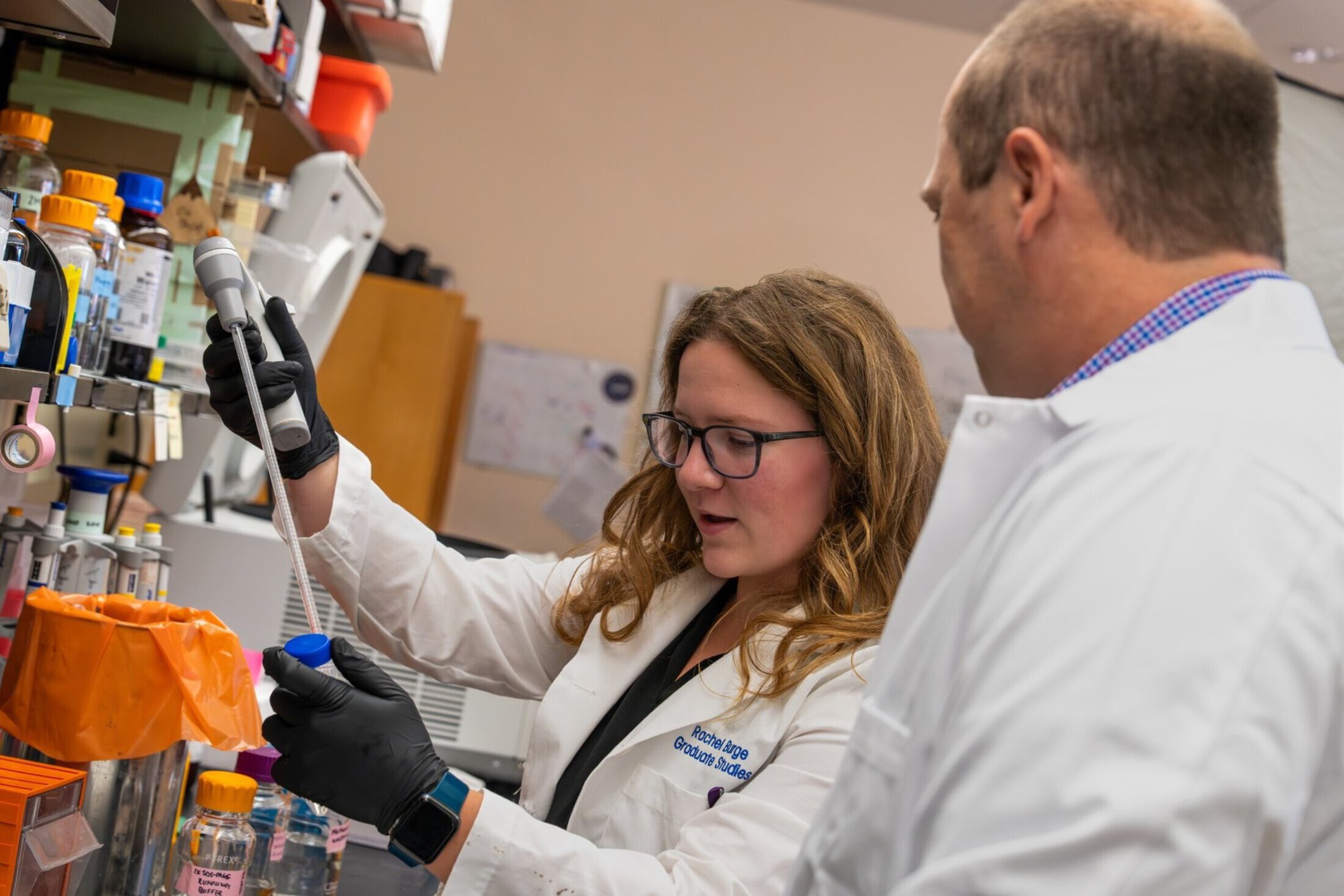 Rachel Burge works in a research laboratory, using a pipette to transfer liquid into a sample tube while a colleague looks on. Laboratory equipment and supplies line the workspace as she conducts pancreatic cancer research.